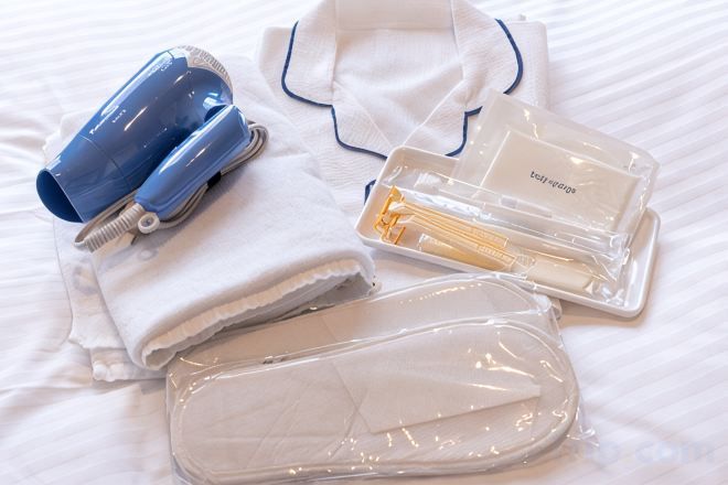 Ein ordentlich arrangiertes Set an Pflegeprodukten auf einem Bett, darunter ein Föhn, eine Lotionflasche und eine Kosmetiktasche.
