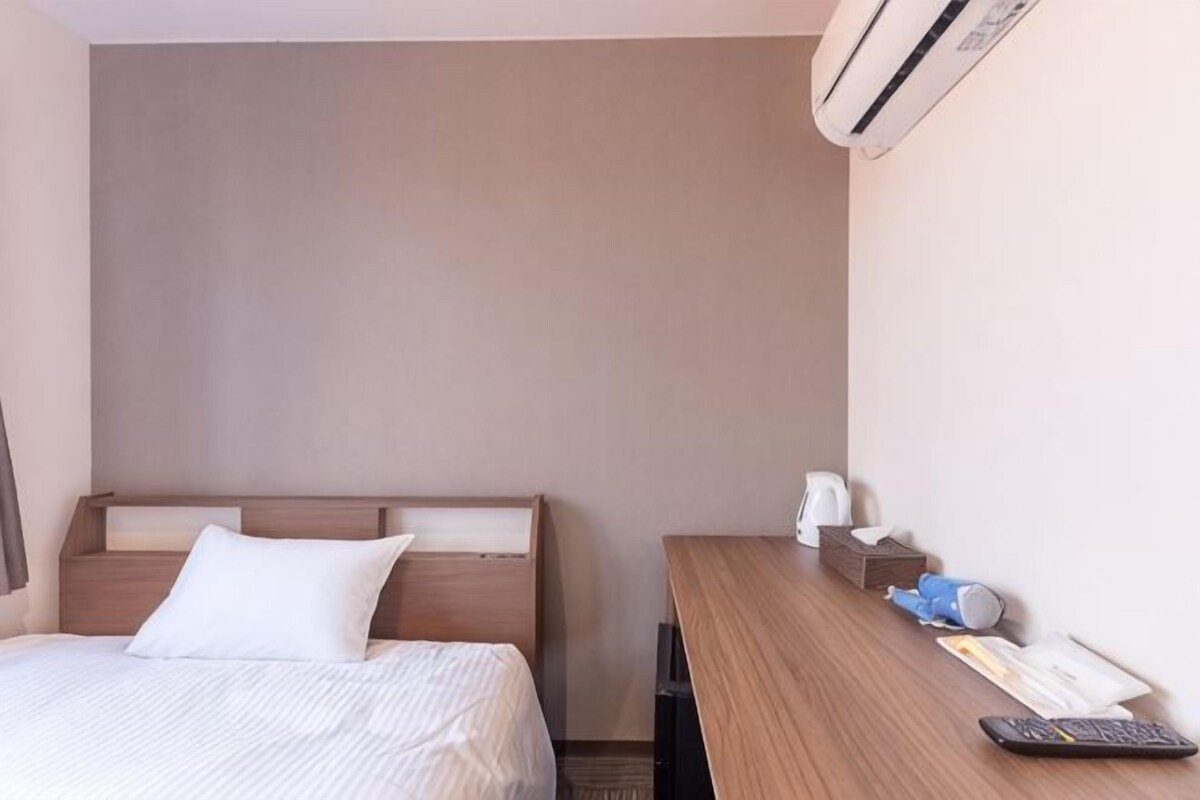 A hotel room with a bed, a desk, a chair, a bedside table, a remote control, and a wall-mounted air conditioner.