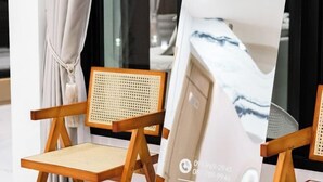 Μια ξύλινη καρέκλα με πλεκτή πλάτη και μαλακό κάθισμα, τοποθετημένη μπροστά από ένα παράθυρο με λευκές κουρτίνες.