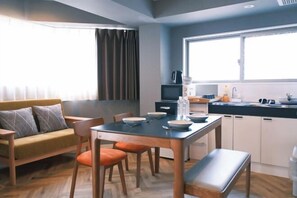 A modern kitchen with a dining table, chairs, and a bench. There is a sofa with cushions, a window with curtains, and a kitchen counter with appliances. - Gracia Hotel (Osaka)