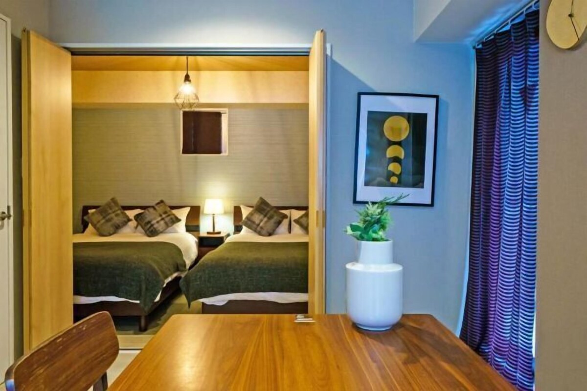 A hotel room with a wooden dining table, two beds with green bedding, a bedside lamp, a framed artwork, and a potted plant.