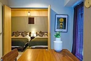 A hotel room with a wooden dining table, two beds with green bedding, a bedside lamp, a framed artwork, and a potted plant. - Gracia Hotel (Osaka)