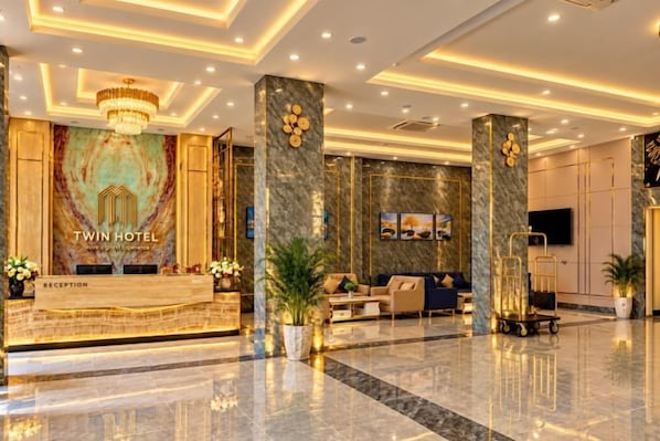 A hotel reception area with a reception desk, a chandelier, a sofa, and a television screen.
