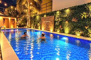 An indoor swimming pool with two people in the water, surrounded by a decorative wall with plants and lights.