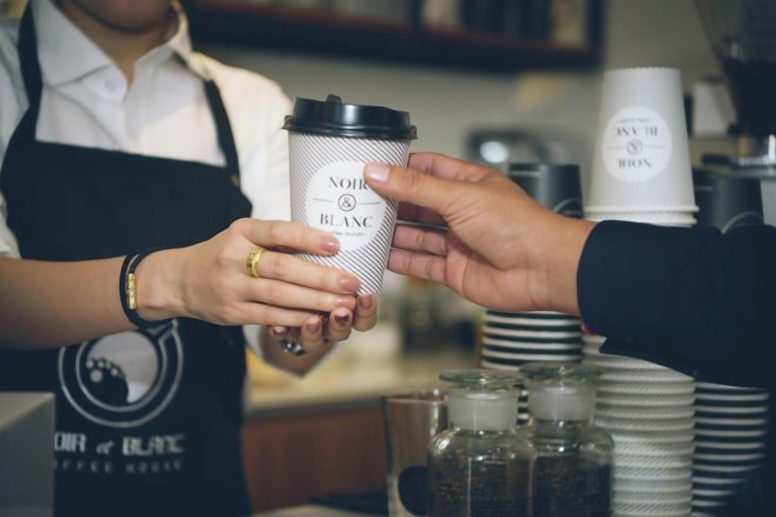 Una persona sosteniendo una taza de café con tapa, otra persona recibiéndola, y una pila de tazas similares al fondo.