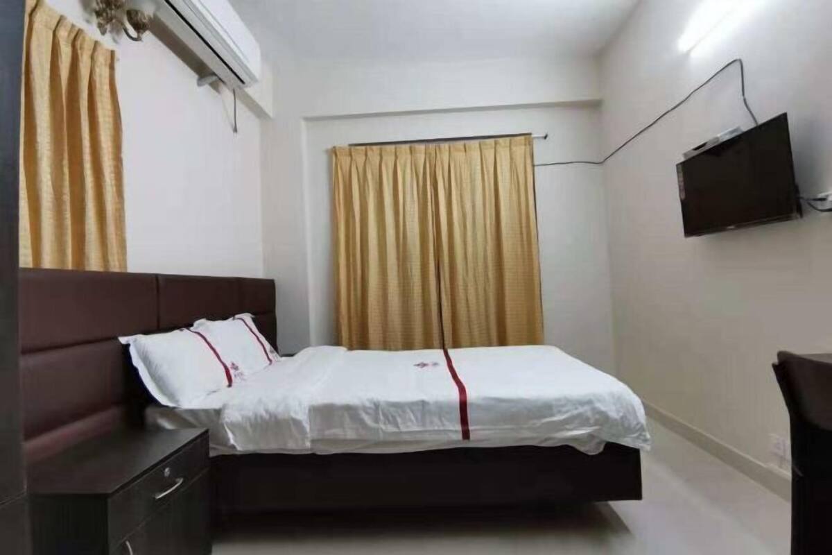 A single bed with a white mattress and red trim, a dark headboard, a bedside table with drawers, a wall-mounted TV, and a window with beige curtains.