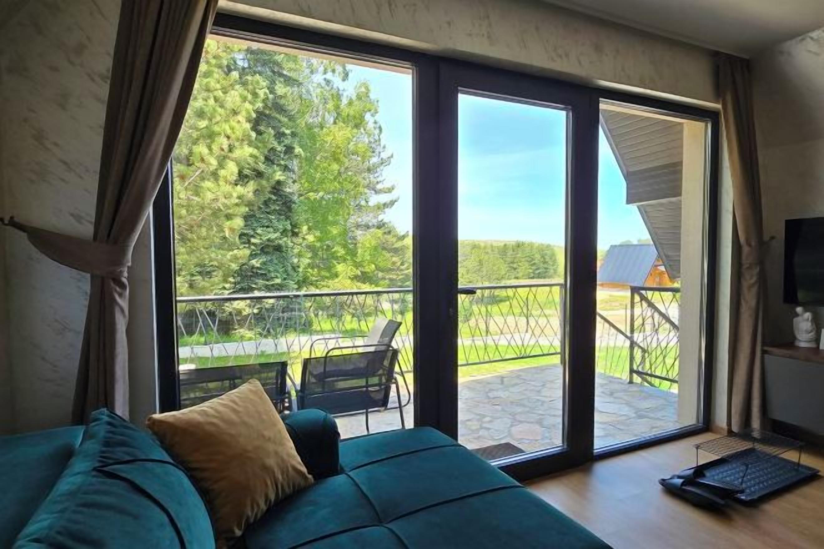 A room with a green sofa, a laptop, and a sliding glass door leading to a balcony with a view of trees and a building.
