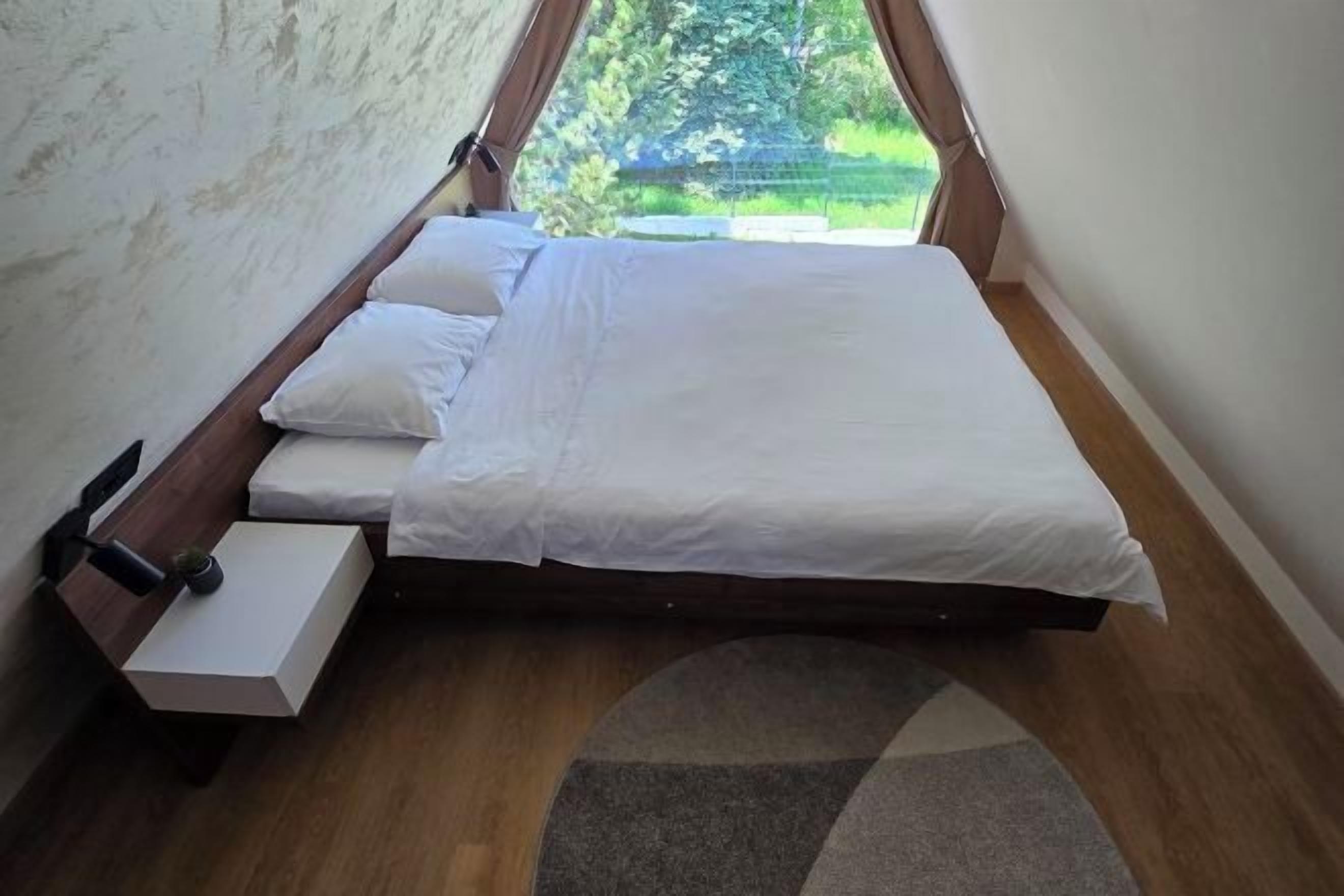 A single bed with white bedding in a room with a sloped ceiling and a view of greenery outside.