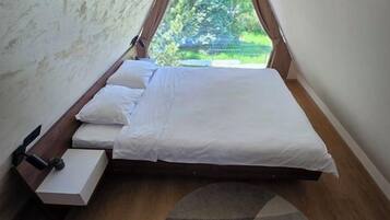 A single bed with white bedding in a room with a sloped ceiling and a view of greenery outside.