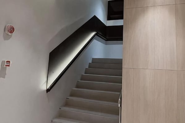 A staircase with a black handrail and white steps, leading to a landing with a light switch and a smoke detector. - Homitel Bangkok (Bangkok)