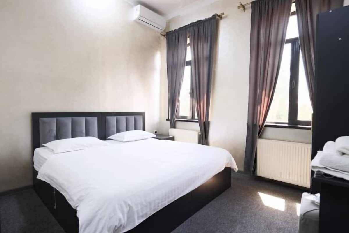 A hotel room with a bed, a nightstand, an air conditioning unit, a window with curtains, and a radiator.