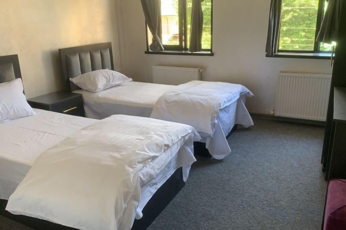 A hotel room with two single beds, a nightstand, a window with curtains, and a radiator.