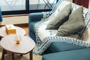 A cozy living room with a blue sofa, a patterned throw blanket, a small wooden table with a glass, and a tissue box. - Garfield Home Shinjuku (Tokyo)