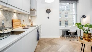 A modern kitchen with white cabinets, a herringbone floor, a hanging light, and a view of buildings through a window. - Kula London - Fitzrovia Newman St (London)