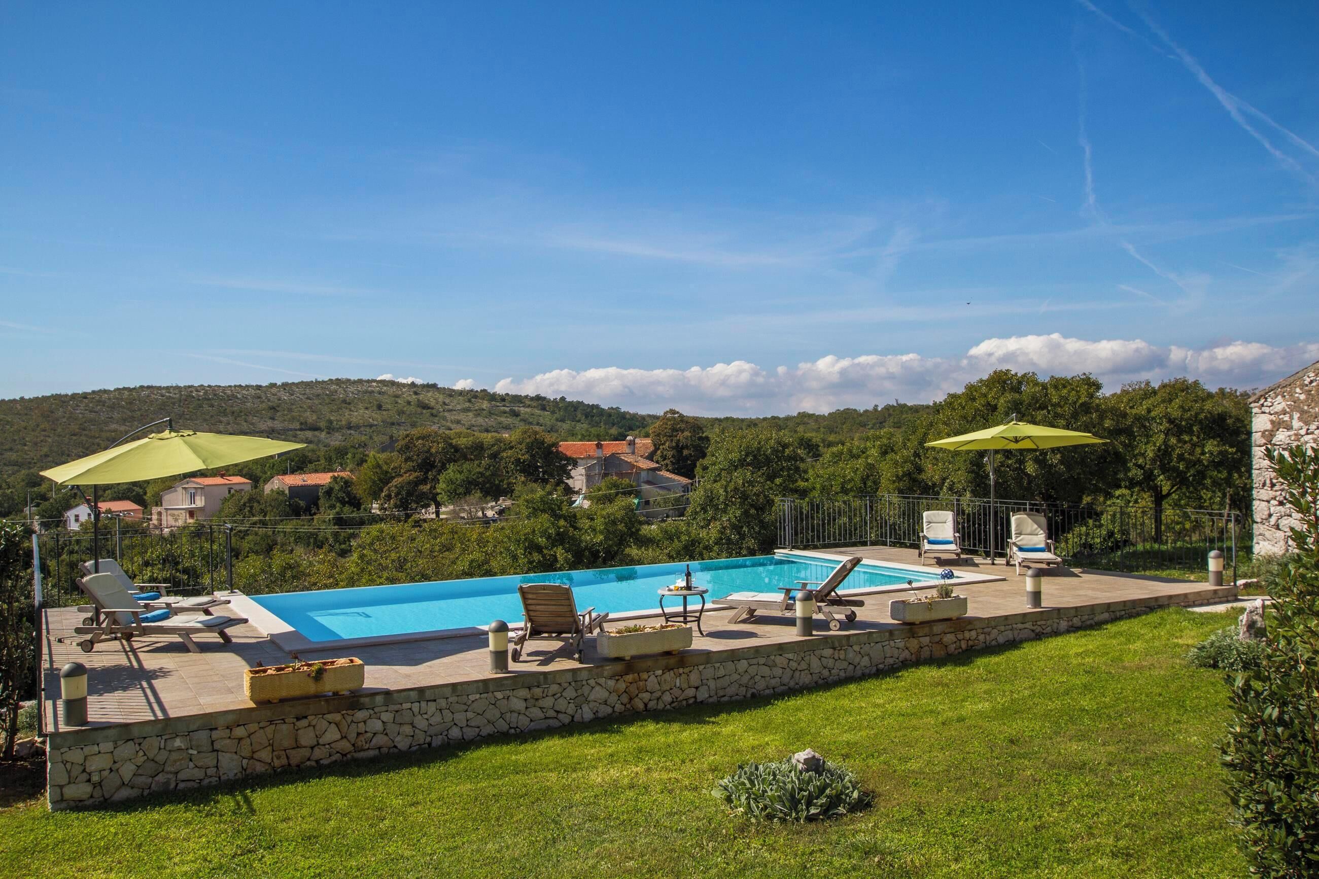 A swimming pool with lounge chairs and umbrellas, surrounded by a stone wall and greenery, with houses and hills in the background.