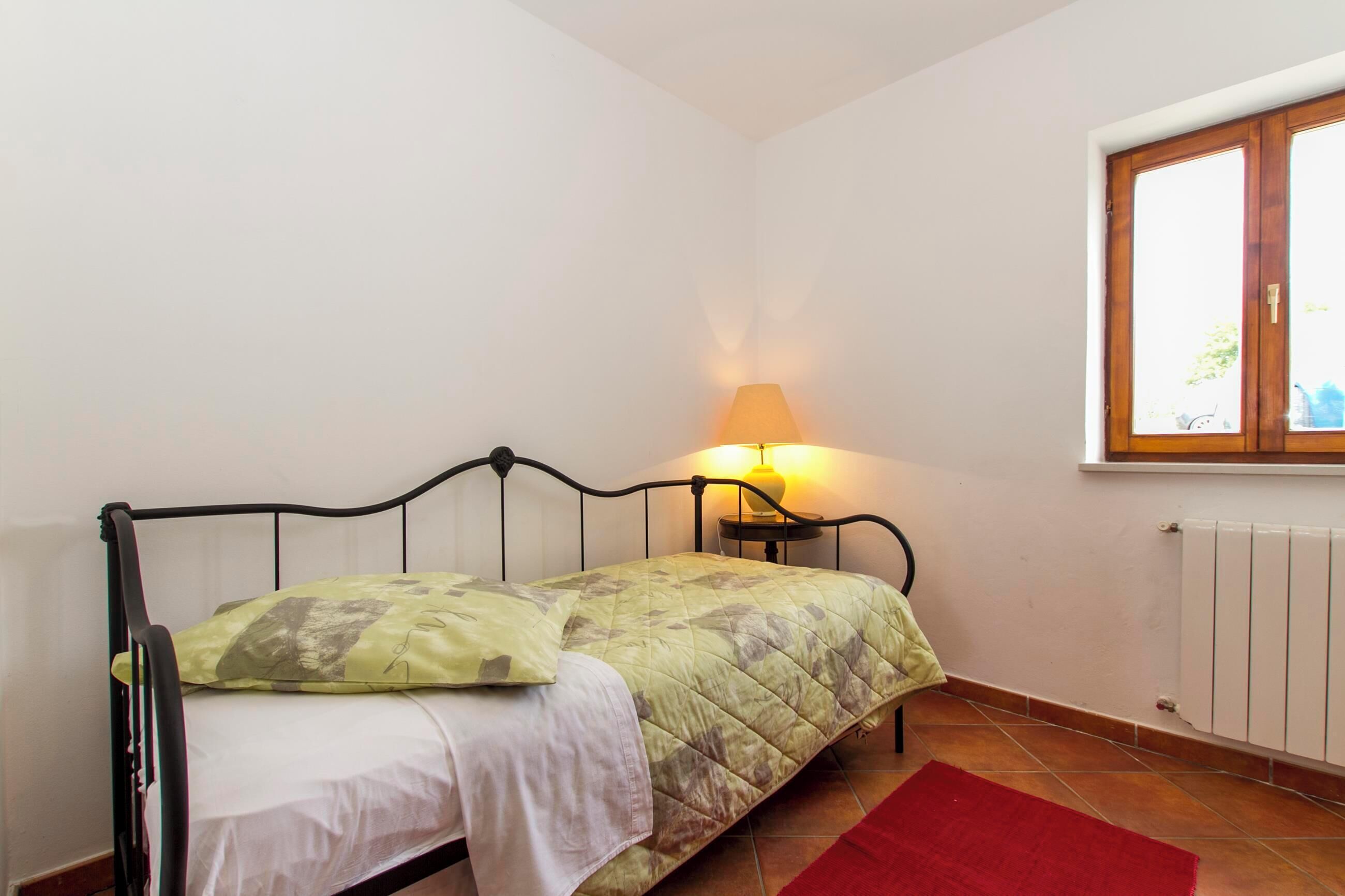 A single bed with a green quilt, a bedside lamp, and a window with blinds.
