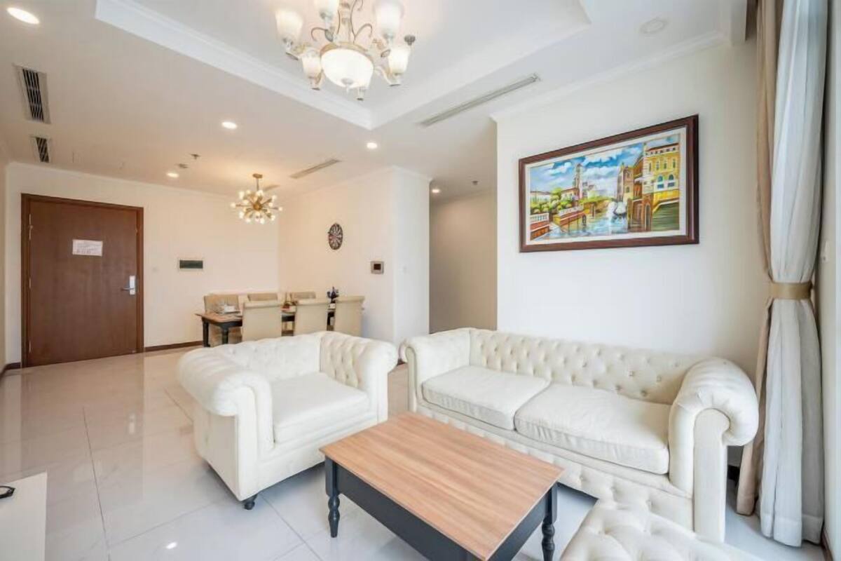 A hotel room with a white sofa set, a wooden coffee table, a chandelier, and a painting of a cityscape.