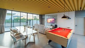 Ein modernes Wohnzimmer mit Billardtisch, einem Esstisch mit Stühlen, einer Couch und einem an der Wand befestigten Fernseher.