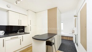 A compact kitchenette with a sink, stove, and cabinets, a small dining area with a table and chair, and a bed in the adjacent room. - Student Only AXO Camden (London)