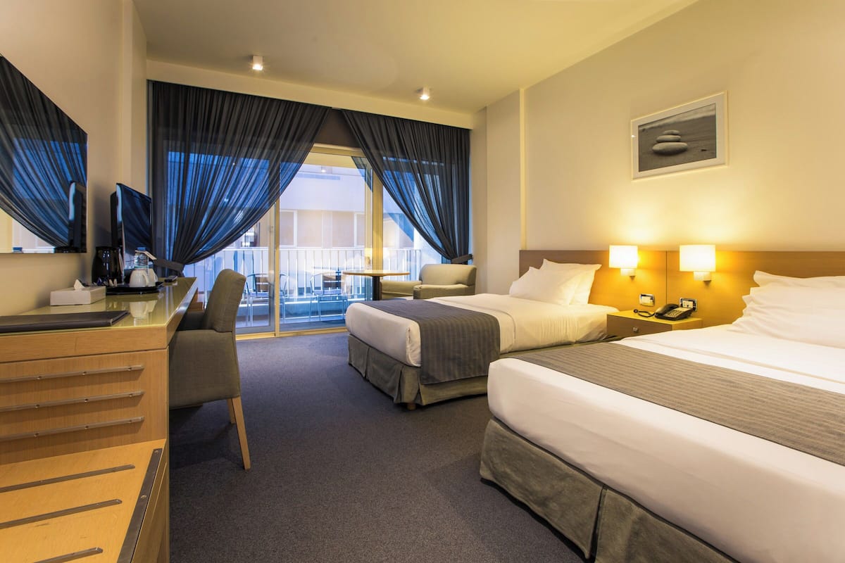 A hotel room with a desk, two beds, a chair, and a balcony with curtains.