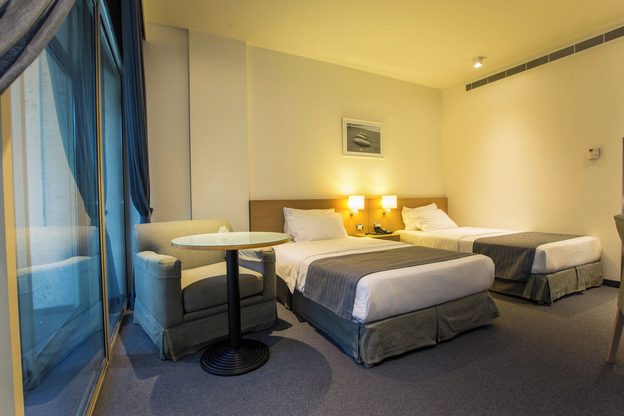 A hotel room with two beds, a small round table, a sofa, and a window with curtains.