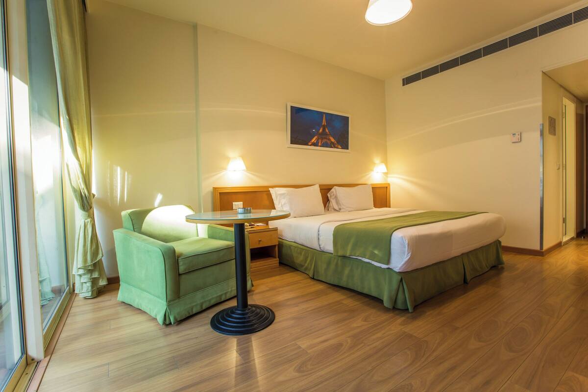 A hotel room with a bed, a green armchair, a small table, and a painting of the Eiffel Tower.