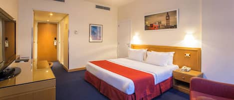 A hotel room with a bed, bedside table, lamp, chair, desk, and a painting of a cityscape.