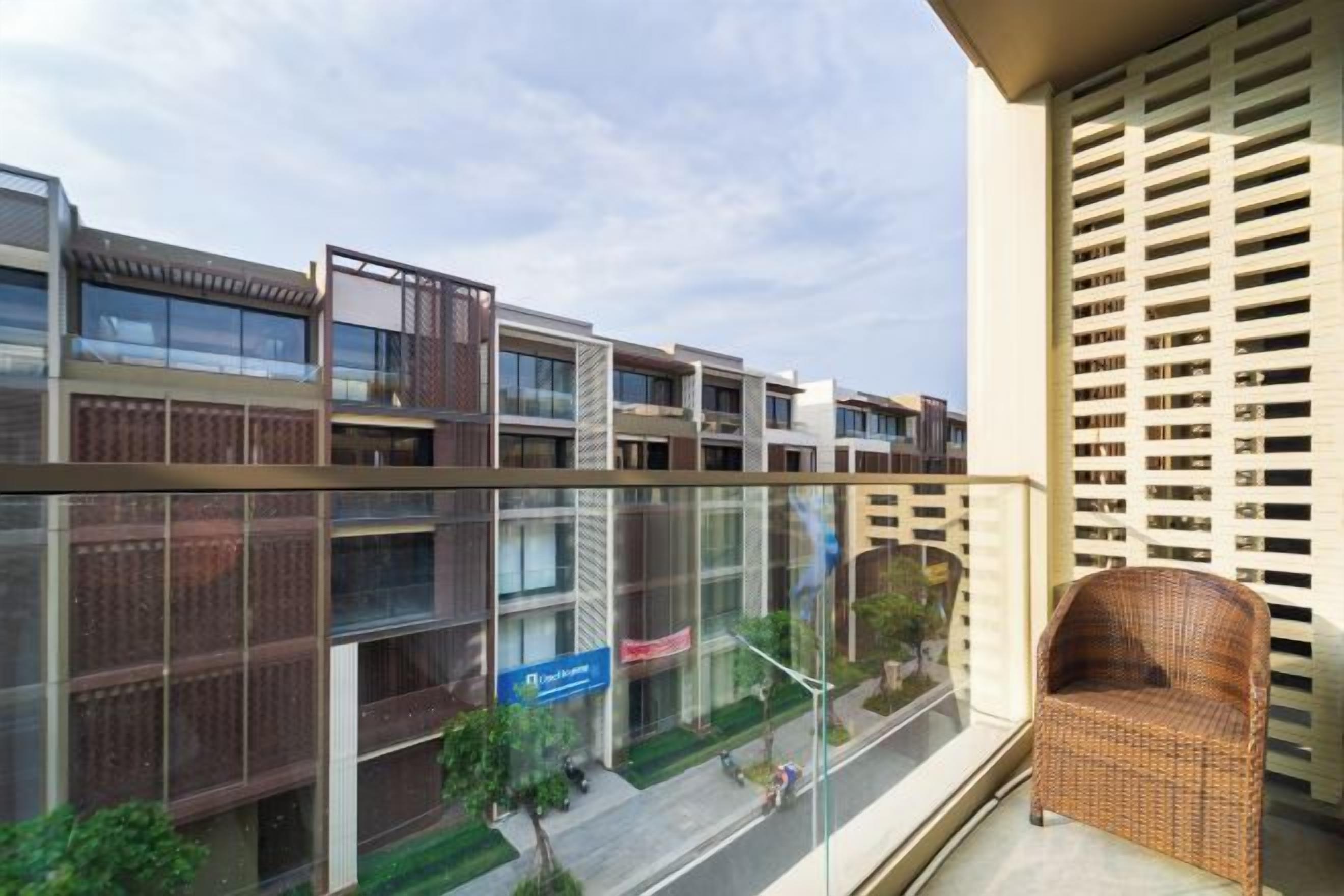 Un balcon avec un fauteuil en osier, donnant sur une rue bordée d’immeubles et un ciel dégagé.