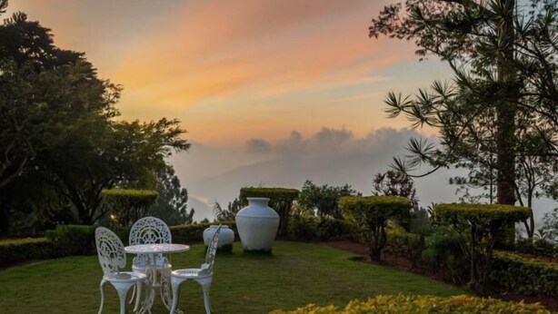 Un jardin avec une table et des chaises blanches, un grand vase blanc et des haies soigneusement taillées.