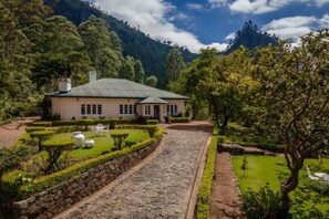 A building with a stone pathway leading to a white structure with a green roof, surrounded by lush greenery and a mountainous backdrop. - Highgrove Estate By Ishq (Nuwara Eliya District)