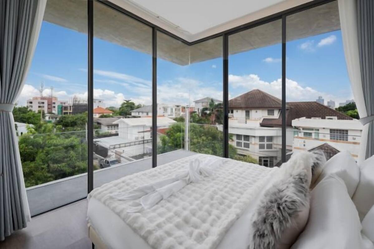 A modern bedroom with a large bed, a view of the city through floor-to-ceiling windows, and a balcony.