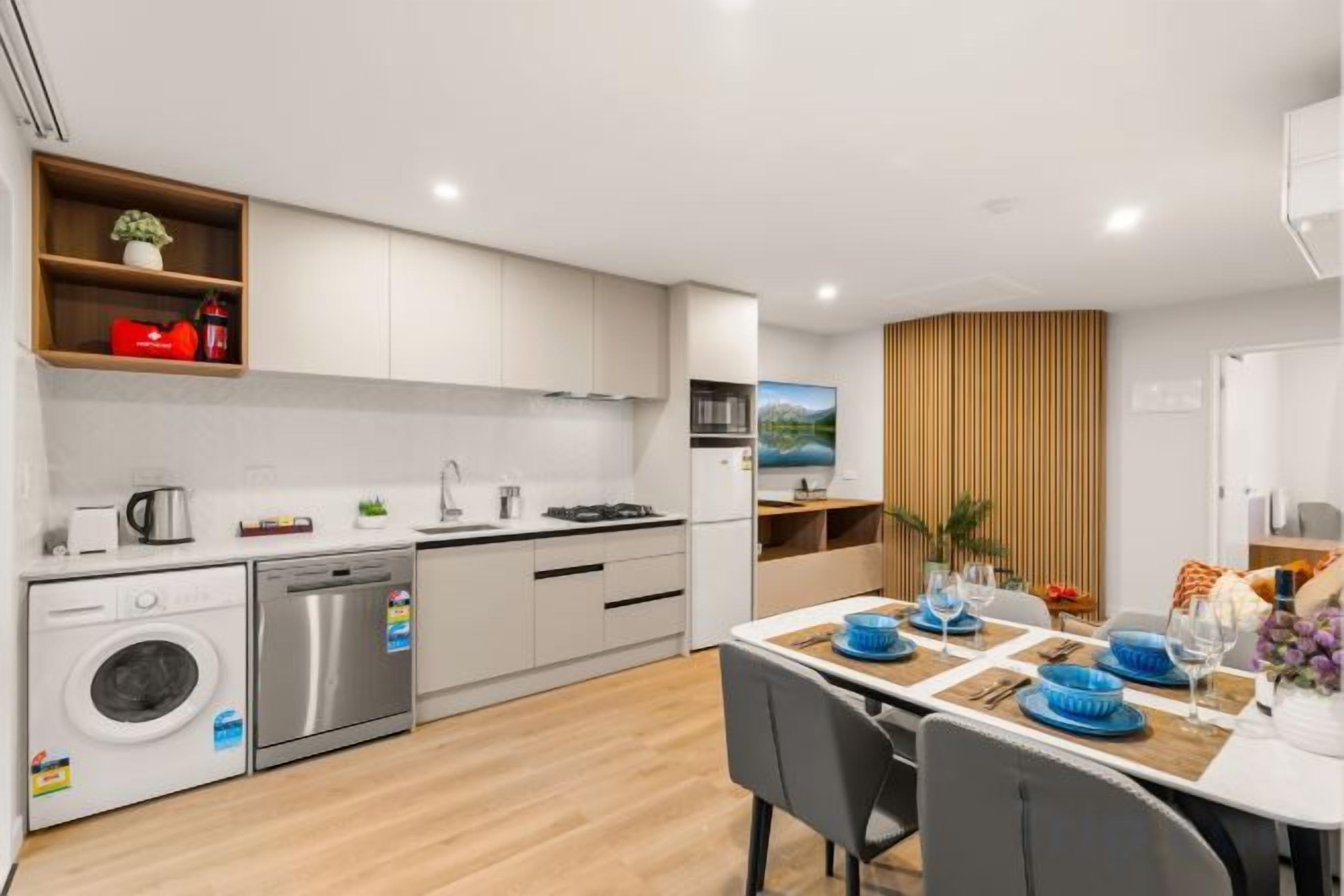 A modern kitchen with white cabinets, a dishwasher, and a sink. There is a dining area with a table set for two, and a window with a view of the outdoors.