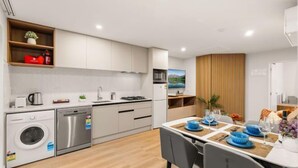 A modern kitchen with white cabinets, a dishwasher, and a washing machine. There is a dining area with a table set for two, blue plates, and glasses. - Starlet 11 (Lake Tekapo)