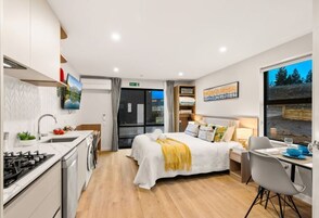 A modern hotel room with a kitchen, a bed with a yellow throw, a dining area, and a large window with a view.