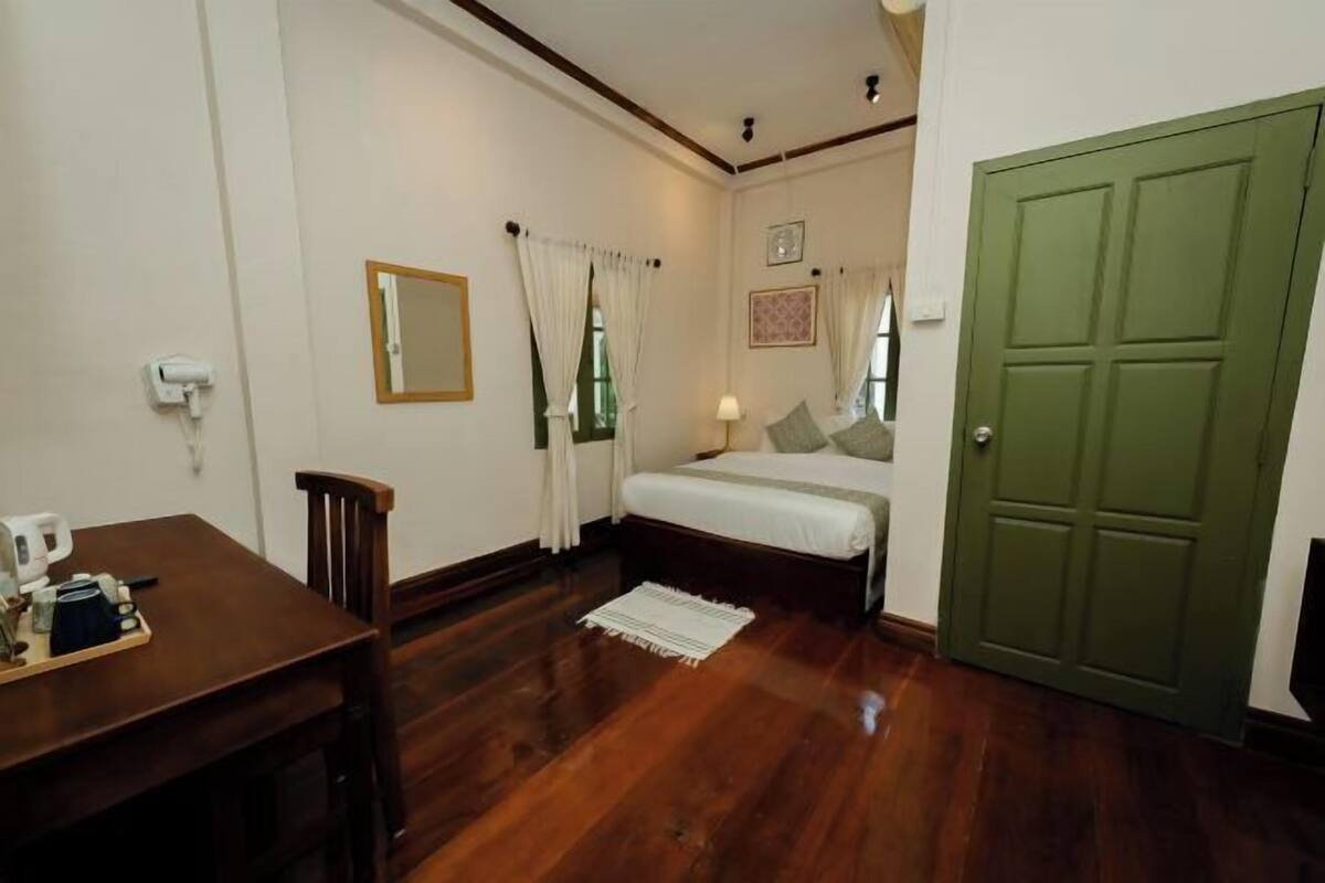 A bedroom with a wooden floor, a bed with white bedding, a green door, and a small table with a coffee maker.