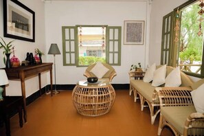 A room with a round wicker chair, a sofa with cushions, a table with a bowl, and a window with green shutters. - Barn Laos Luangprabang Heritage (Luang Prabang)