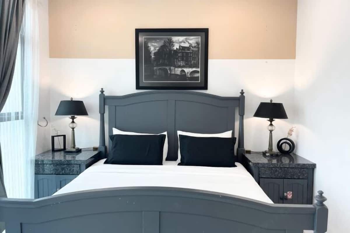 Une chambre à coucher bien rangée, avec un lit placé au centre, entre deux tables de chevet, chacune surmontée d’une lampe. Un tableau encadré est accroché au-dessus du lit.