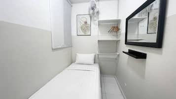 Quarto pequeno, branco, com uma cama de casal, parede branca, espelho com moldura preta e um ventilador branco.