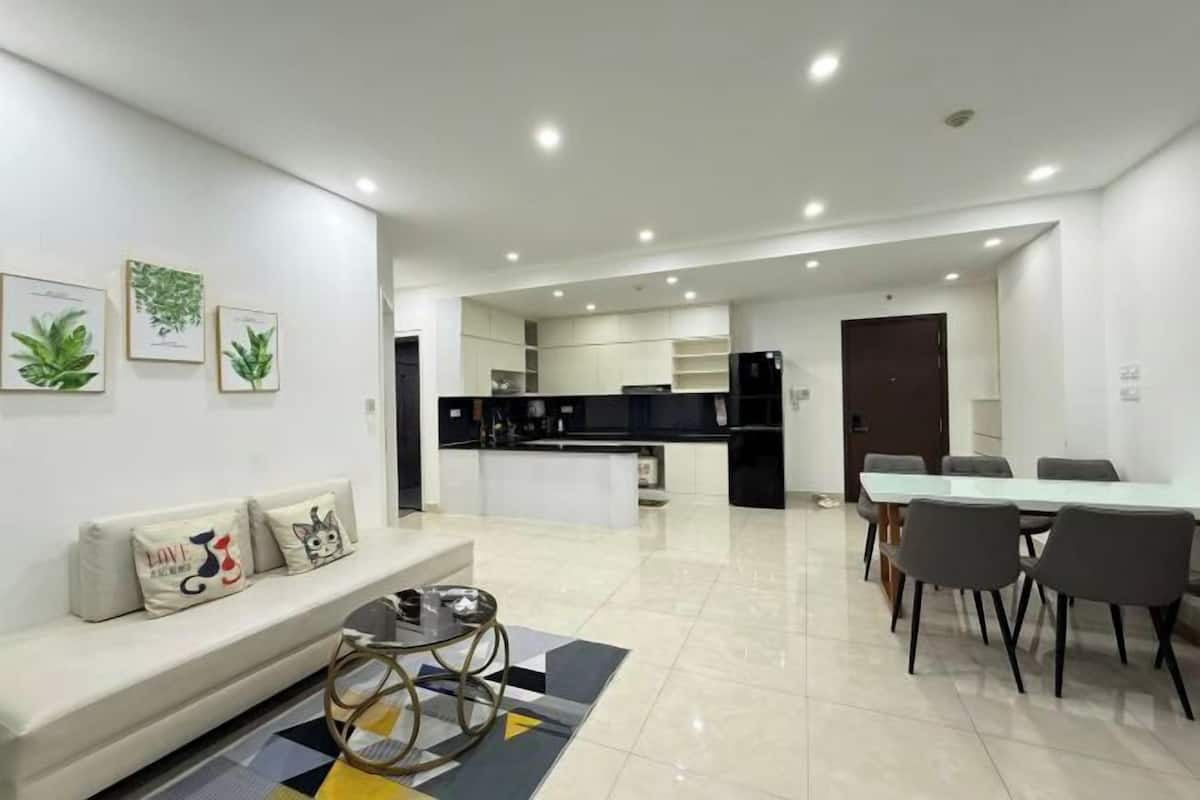 Ein modernes Wohnzimmer mit einer Couch, einem Esstisch und Stühlen. Im Hintergrund ist eine Küchenzeile mit Schränken und einem Kühlschrank zu sehen.