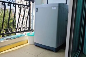 A balcony with a metal railing, a large gray appliance, and a view of greenery. - City Garden Olympus Condominium 1room (Pattaya)