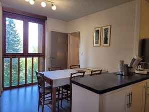 Uma sala de jantar com mesa e cadeiras, bancada de cozinha com eletrodomésticos e janela com vista para árvores.