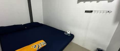 A small, single-bed room with a blue mattress, a towel, and a sports jersey. There is a window with curtains, a wall-mounted rack, and a trash bin.