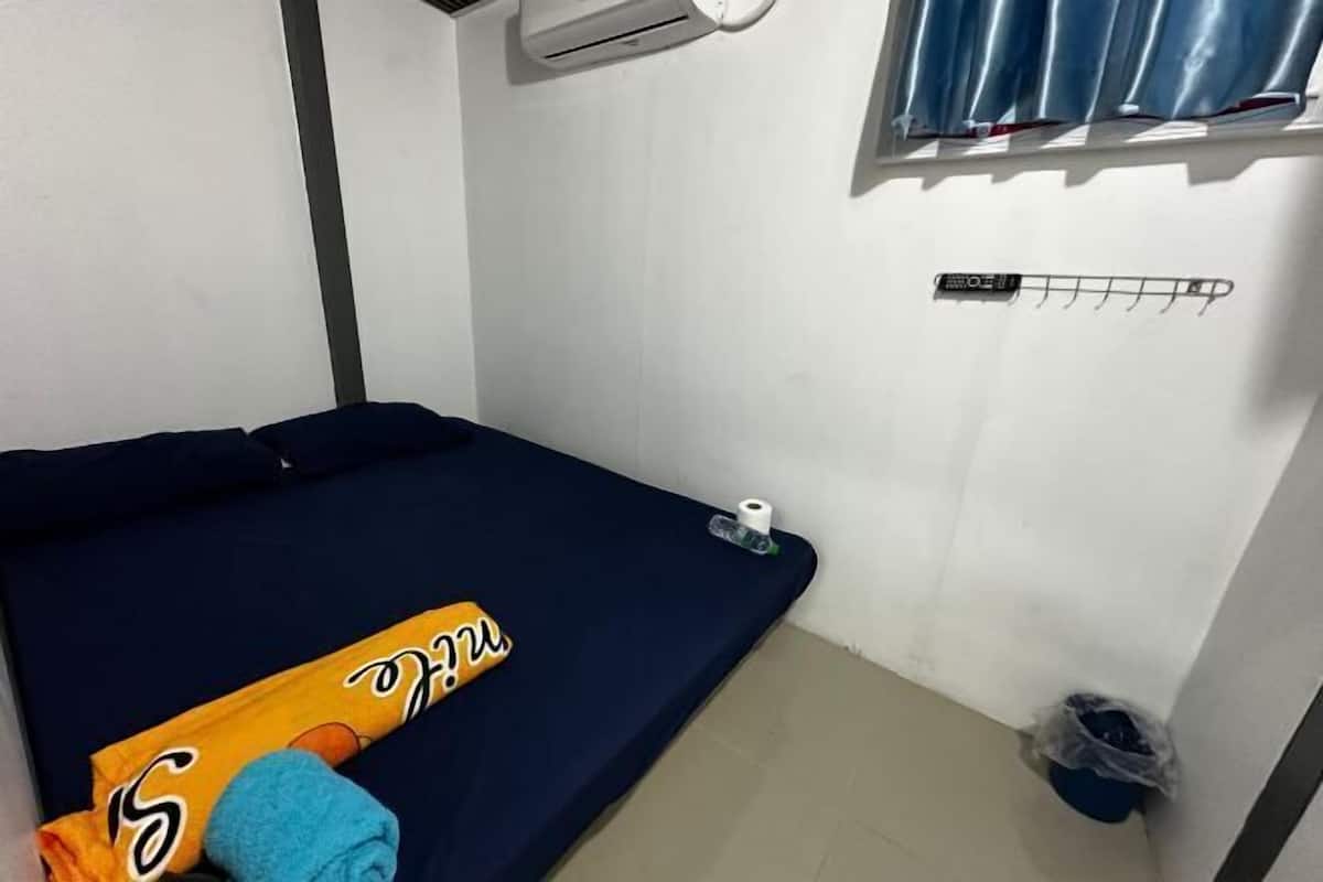 A small room with a bed, a window with curtains, a wall-mounted air conditioner, a towel, and a trash bin.