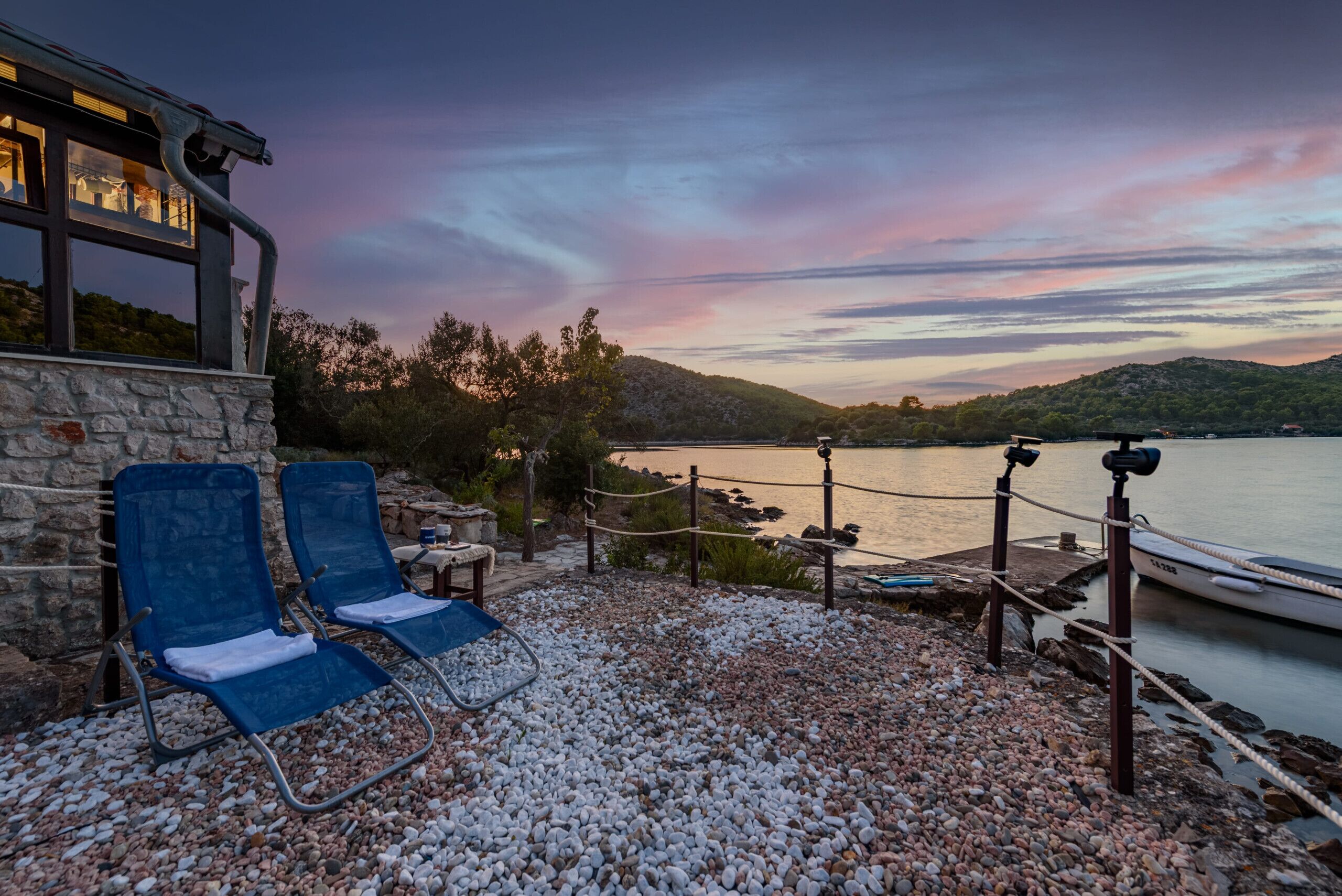 Una terraza junto al lago con dos sillones azules, un muro de piedra y vistas al lago y las colinas al atardecer.