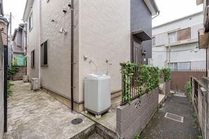 A narrow alleyway with a white building on the left, a water tank, and a small garden area with a fence. - W.Hostel (Tokyo)