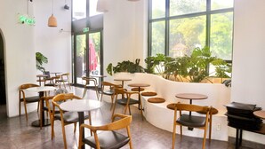 A cafe with round tables and wooden chairs, large windows with a view of greenery, and indoor plants. - Monstera Hotel and Coffee (Phu Quoc)
