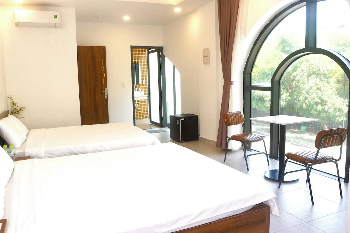 A hotel room with two beds, a small table with a chair, and a large window offering a view of greenery.
