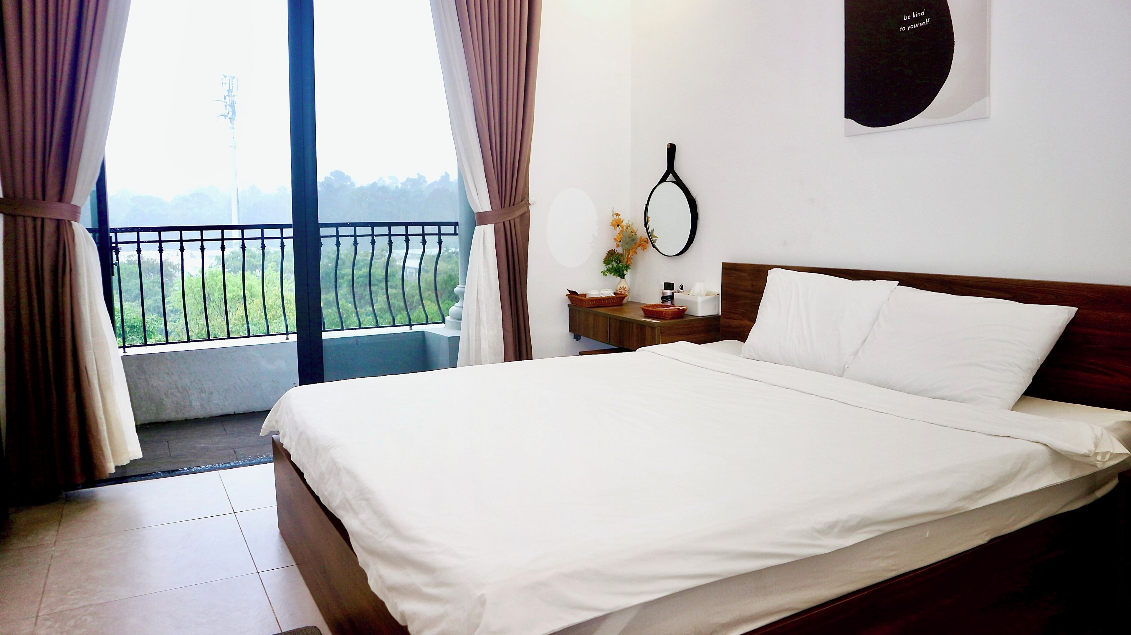 A hotel room with a large bed, a nightstand with a lamp, a mirror, and a view of greenery outside.