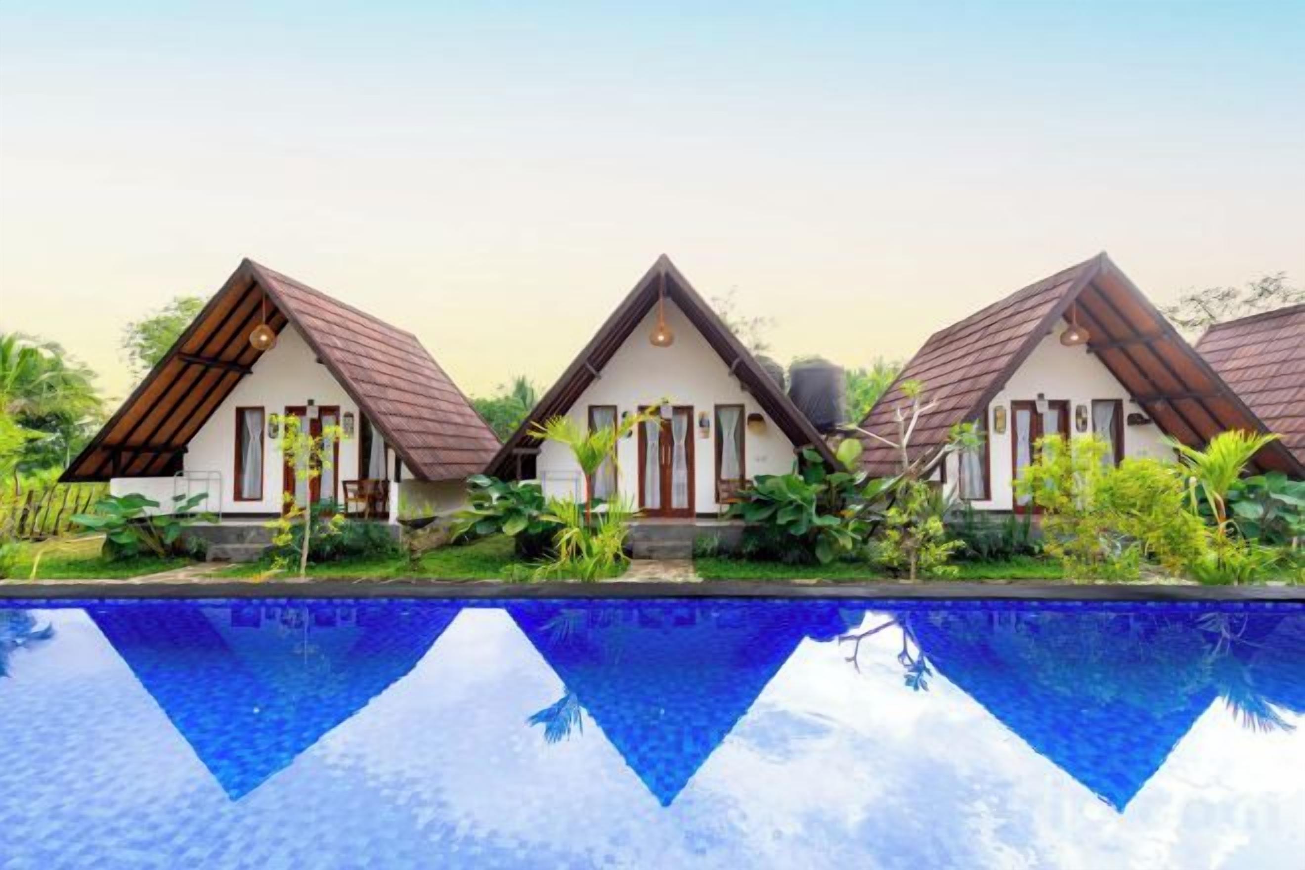 Three bungalow-style buildings with pitched roofs, a swimming pool in the foreground, and lush greenery surrounding the property.