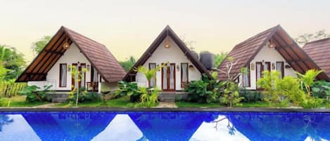 Three bungalow-style buildings with pitched roofs, a swimming pool in the foreground, and lush greenery surrounding the property.